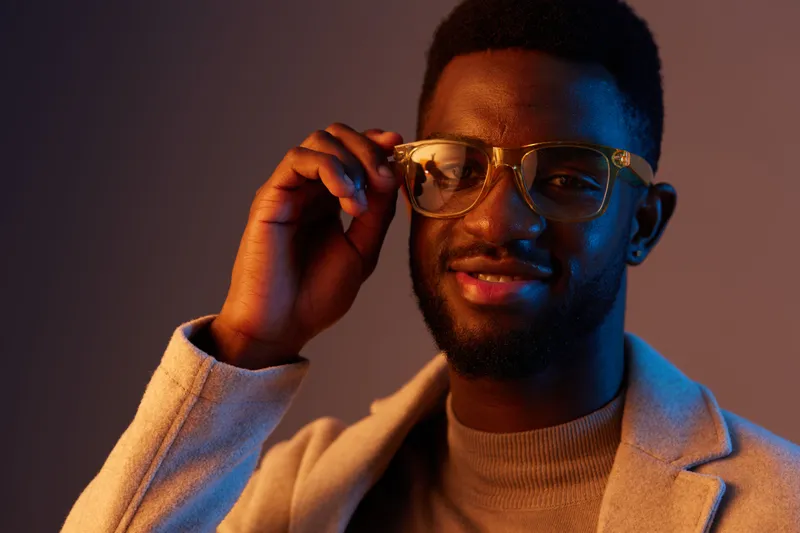 Confident young Black man wearing stylish glasses and a light jacket with a warm colored background, showcasing a modern and trendy atmosphere