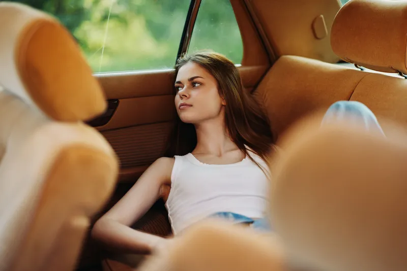Young woman relaxing in a car, wearing a white tank top and casual jeans, expressing a serene emotion in a warm, natural background.