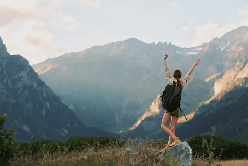 A mountain hike unfolds as a young woman hiker with a backpack raises her arms in triumph on a rocky ledge, framed by alpine scenery under a clear sky.