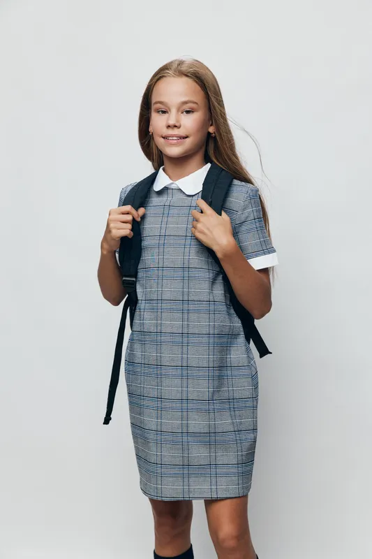 Girl with backpack wearing a checkered dress standing on white background, smiling and posing for school or education concept with confidence and youth style.