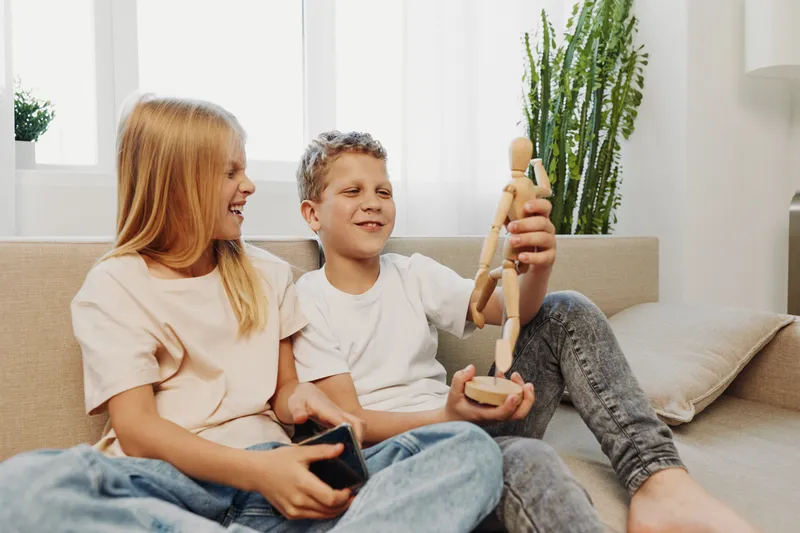 Happy children sitting on sofa at home, boy holding wooden mannequin and smiling while girl watches with joy. Cozy bright living room with natural light and indoor plants.