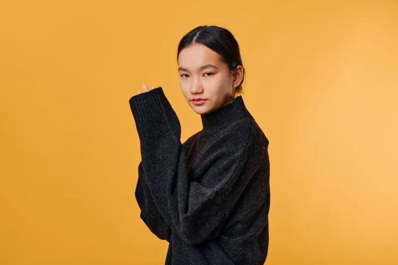 Young woman in black sweater posing with crossed arms and calm expression against orange background, fashion portrait with casual style and natural makeup for modern look.