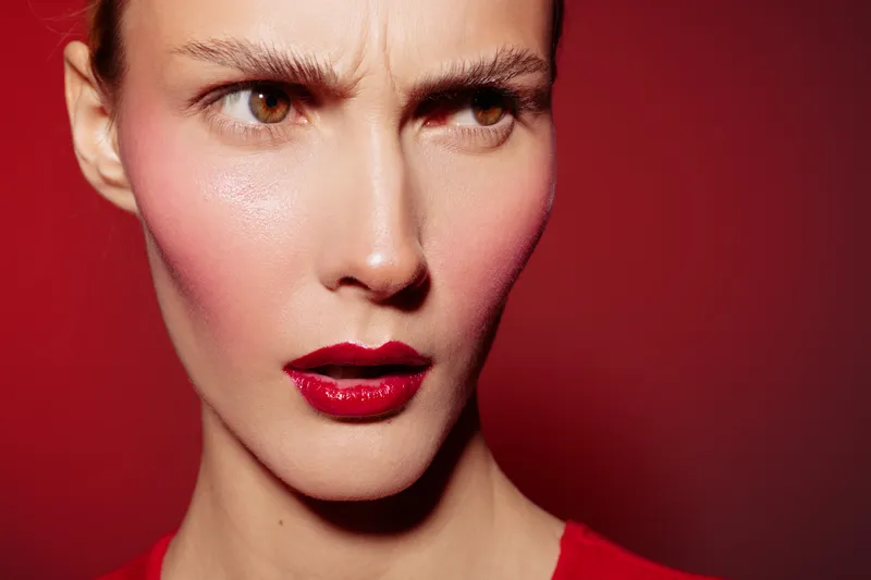 Close-up of a young woman with red lipstick and furrowed brows against a red background. Concept of emotion, beauty, makeup, and intense facial expression in studio lighting.