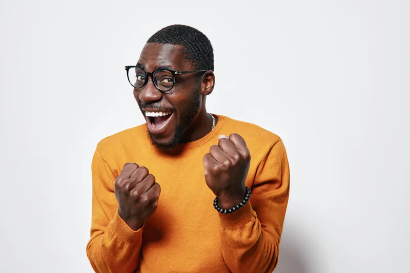 African American man with dark skin wearing glasses and orange sweater smiling excitedly with clenched fists on white background. Happy, joyful, casual portrait of young black male.