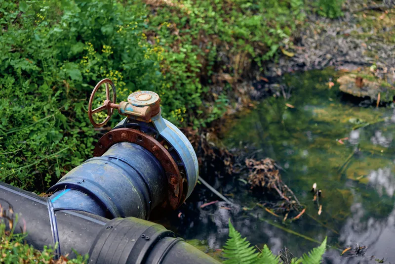 Pipe valve and water near rusted flange with visible leak at pond edge, industrial pipe discharging into shallow pond among vegetation, wetland contamination and muddy bank detail.
