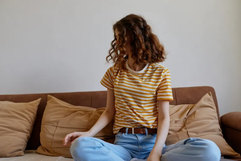 Young woman sits cross legged on a sofa at home, wearing a striped yellow t shirt and light blue jeans, looking away with a calm, reflective mood.