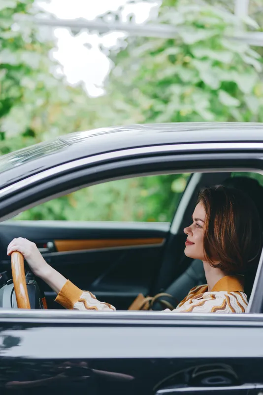 A woman drives a sleek car, focus on her calm profile behind the steering wheel as she glides along a sunlit road.