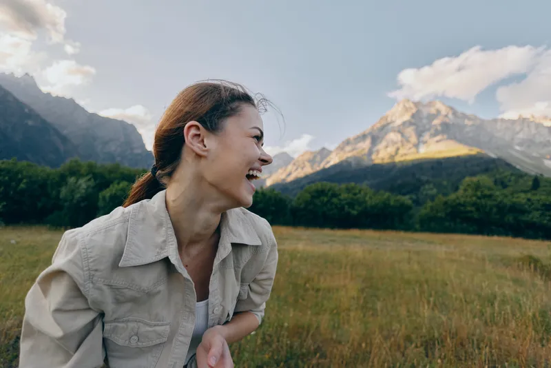 Smiling woman outdoors in a field with mountains in the background. Casual travel lifestyle and natural light create a carefree, vibrant mood that inspires wellness.