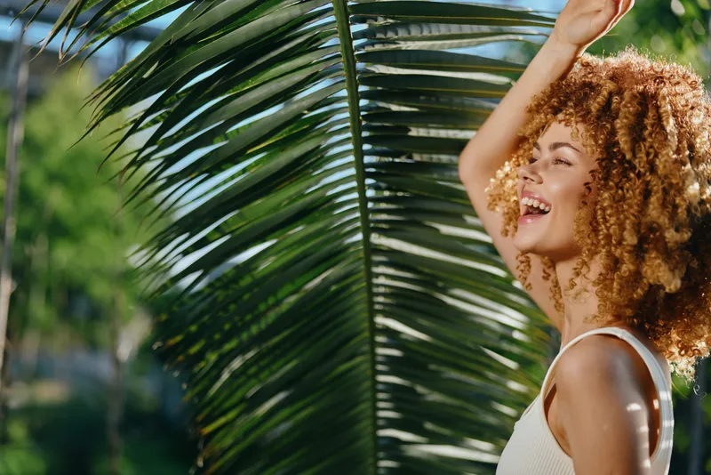 smiling woman with curly hair enjoying sunny tropical outdoors near palm leaves, wearing white tank top and expressing happiness and freedom under bright daylight.