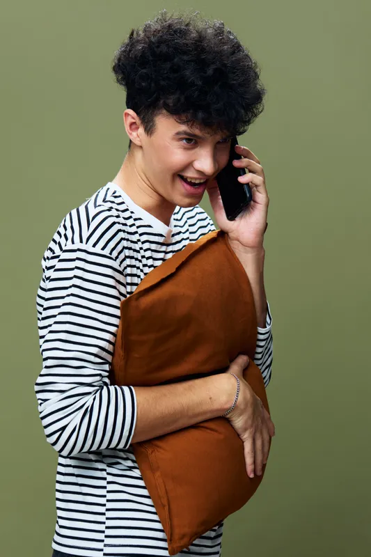 smiling young man talking on smartphone with striped shirt holding brown pillow, casual indoor portrait showing relaxed joyful expression, olive green background