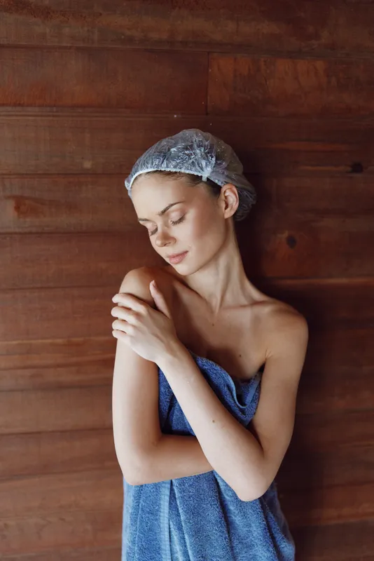 Young woman wrapped in towel with shower cap stands relaxed against wooden wall, displaying peaceful and fresh vibe with natural beauty and soft light.