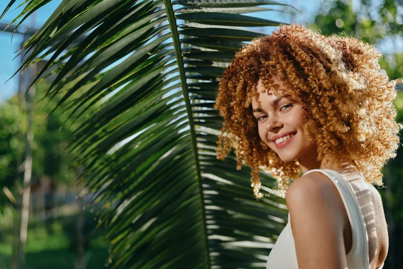 smiling woman with curly hair stands near palm leaves outdoors, warm sunlight highlights her natural beauty and joyful expression in summer atmosphere
