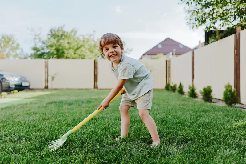 Happy boy playing in the garden, holding a rake and smiling brightly. The scene captures joy and curiosity in a vibrant outdoor setting.