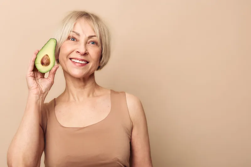 Smiling middle aged woman with short blonde hair holds half of fresh avocado close to face on beige background. Concept of healthy lifestyle, natural food, nutrition, and wellness.