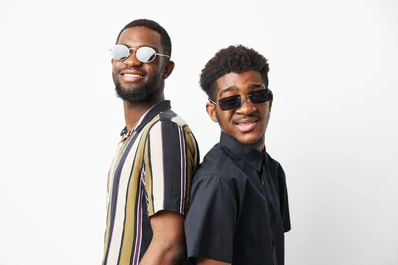 Two stylish African American men wearing sunglasses stand back-to-back, exuding confidence and friendship. Their outfits are trendy and casual, perfect for a fun summer day.
