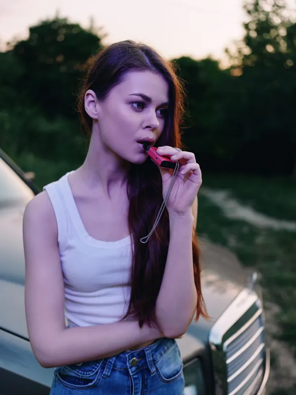 Young woman with long hair holding a whistle, standing beside a car in an outdoor setting during sunset, conveying determination and focus.