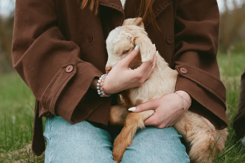 A young woman in a brown jacket gently holds a baby goat on her lap outdoors, showcasing a tender moment of connection and care in a natural lifestyle setting.