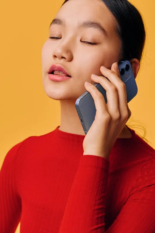 Young woman with closed eyes talking on mobile phone indoors against yellow background. Portrait of female with long black hair wearing red sweater using smartphone for conversation.