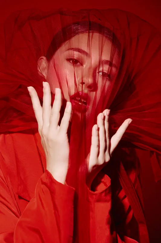 Woman in dramatic red sheer fabric create vibrant fashion portrait. Her lips glossy red, hands gently framing face. Concept of beauty creativity and avantgarde style ensemble.