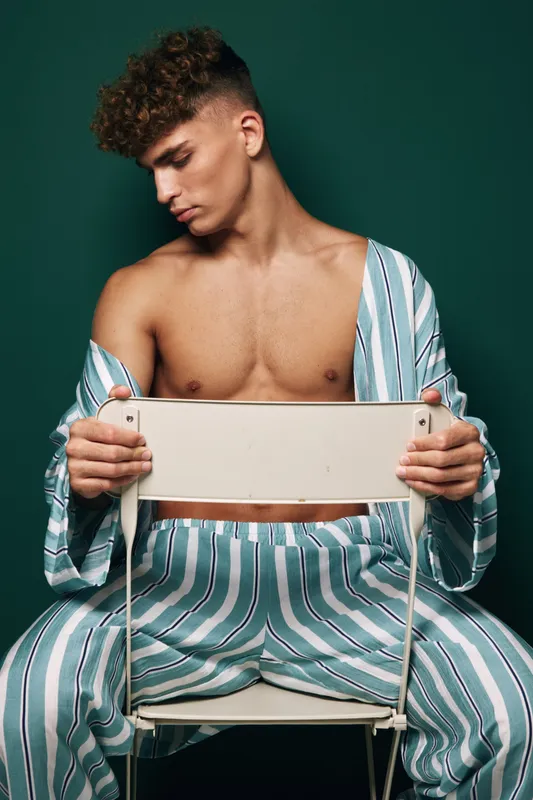 Young man wearing striped shirt and pants sitting on chair backwards looking down in contemplation. Male model with close cropped hair, bare chest, and relaxed posture in studio environment.