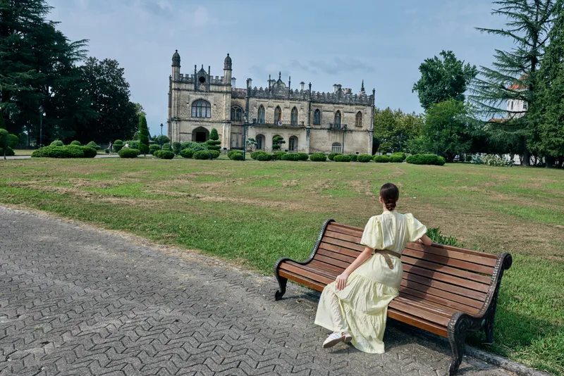 A bench in a park hosts a woman in a light dress, gazing toward a castle, creating a calm landscape and timeless outdoor moment.