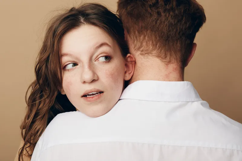 Young woman with red hair embraces man wearing white shirt, looking sideways with thoughtful expression. Intimate moment, relationship, affection, and emotional connection concept.