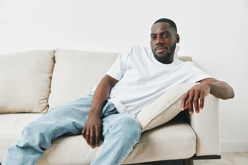 A young African man in a casual white t shirt and jeans, relaxed on a couch, conveying confidence and comfort in a bright, minimalistic setting