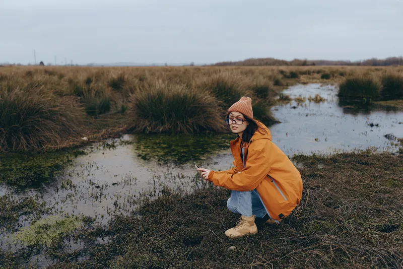 woman lifestyle outdoors in nature, autumn river landscape, orange coat hat walking on muddy trail by wetland grass and water, calm joyful mood on cloudy daylight, with warm light today
