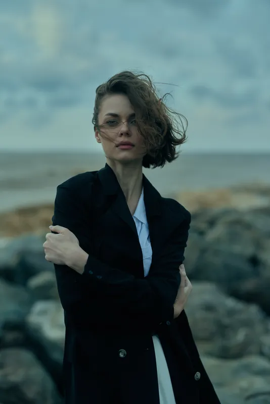 Windswept woman in a dark blazer stands among rocky coastlines, gazing into the distance with a calm, confident expression, wind in her hair, moody sky and sea backdrop, highlighting fashion, outdoor