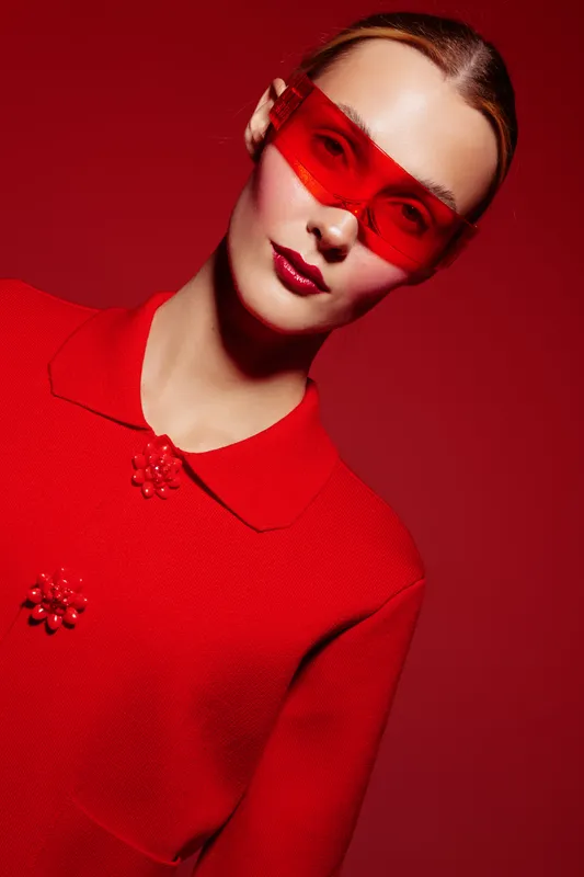 Young woman in red sweater and red translucent glasses poses against red background. Fashion portrait with glossy lips and sleek hairstyle. Concept of style, color, and modern beauty.