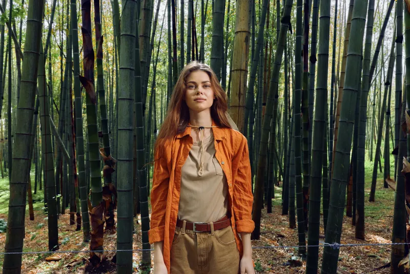 Woman standing in a bamboo forest, portrait of a model in orange jacket and brown pants with long hair. Nature scene with tall trees, green foliage, sunlight and serene outdoor mood.