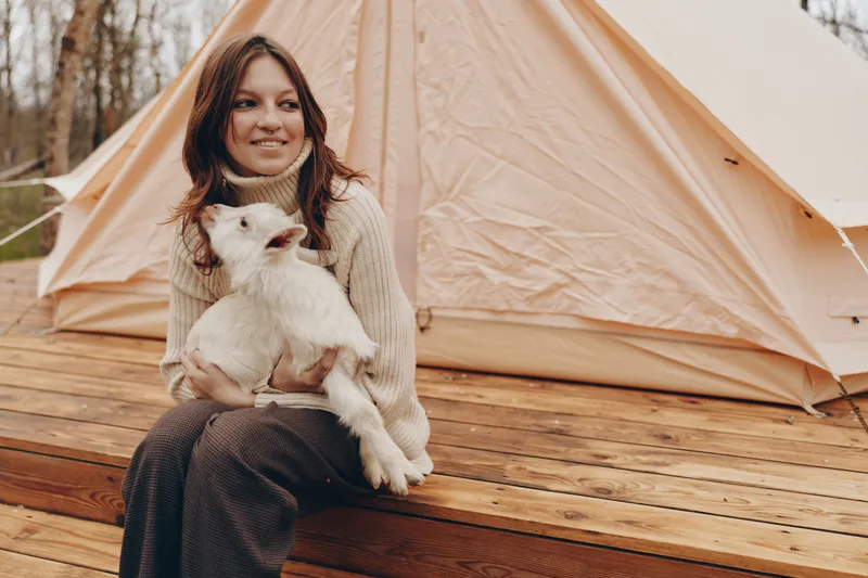 A young woman in a beige turtleneck sweater sits on wooden steps holding a white baby goat near a tent during an outdoor lifestyle camping scene.