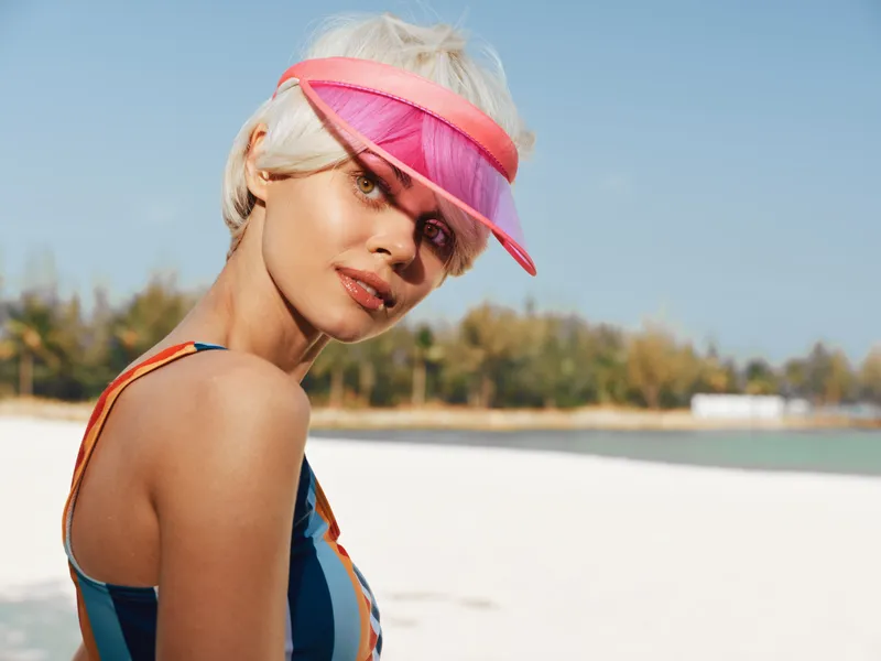 Closeup portrait of a stylish woman at a sunny beach, wearing a bright pink visor and colorful swimsuit, capturing bold fashion and sunlit attitude.