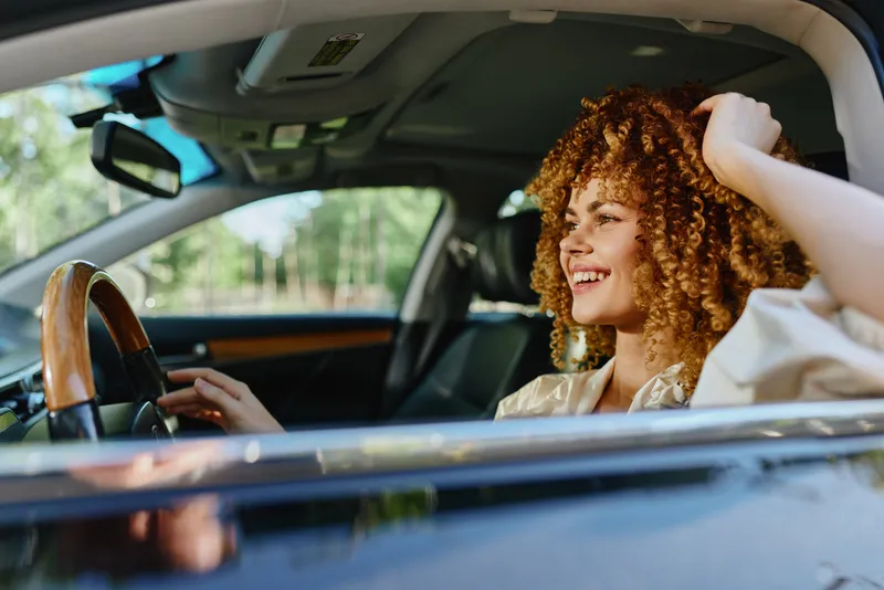 Smiling woman driving a car with curly hair, enjoying a sunny day, relaxed and happy, casual clothing, modern vehicle interior.