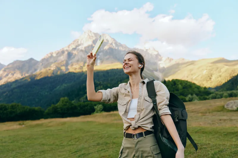 Young woman with a backpack stands in a grassy mountain meadow, holding a map up high and smiling as the sun shines on rugged peaks, signaling adventure