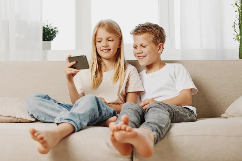 Two happy children, boy and girl, sitting barefoot on a couch, smiling while looking at a smartphone together in a bright living room. Concept of childhood, technology, and friendship.