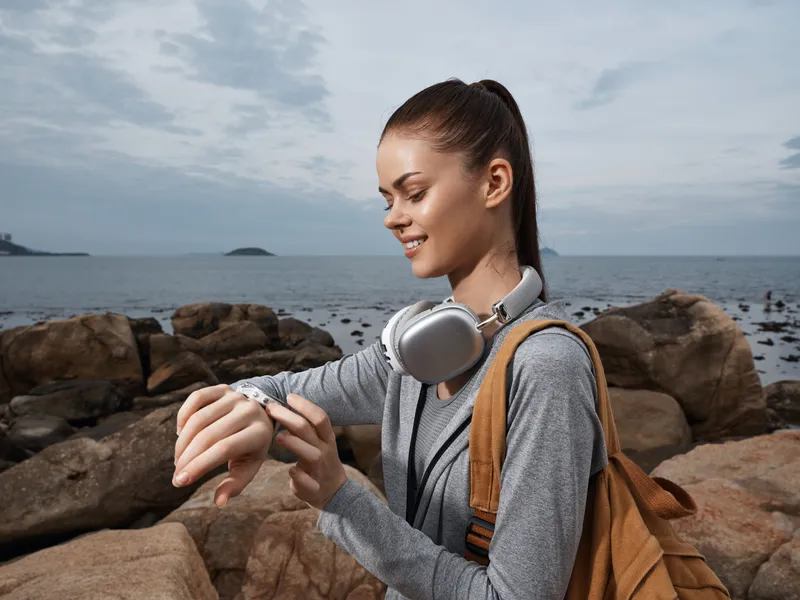 Young woman checking fitness tracker while enjoying a serene day at the beach, wearing casual sportswear and headphones, with a tranquil ocean background