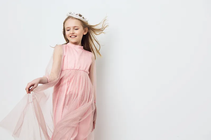 Cheerful girl in a pink dress with flowing fabric and a crown posing against a clean white background, exuding happiness and joy
