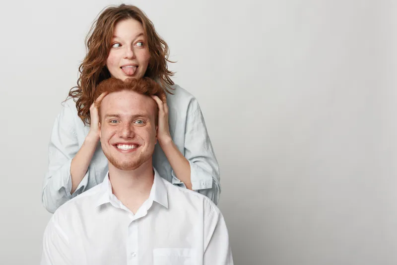 Happy young man with red hair smiles while woman with curly hair playfully covers his eyes and sticks out tongue. Studio portrait on light background. Concept of friendship and fun moments.