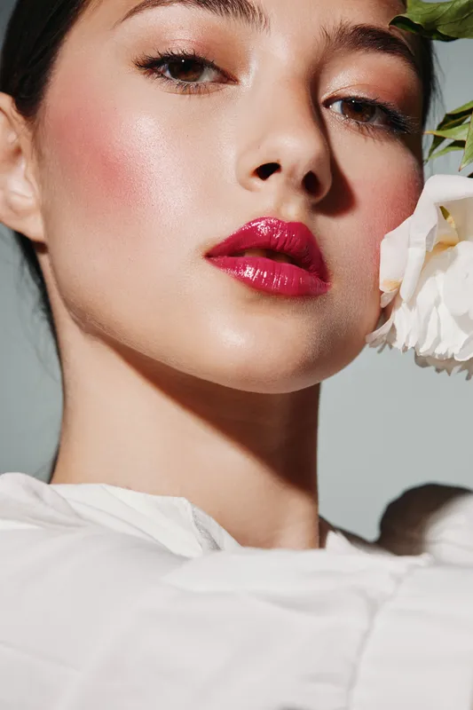 Close-up of a female model with glossy red lips and radiant skin holding a white flower against a neutral background. Beauty and fashion creative in soft natural light.