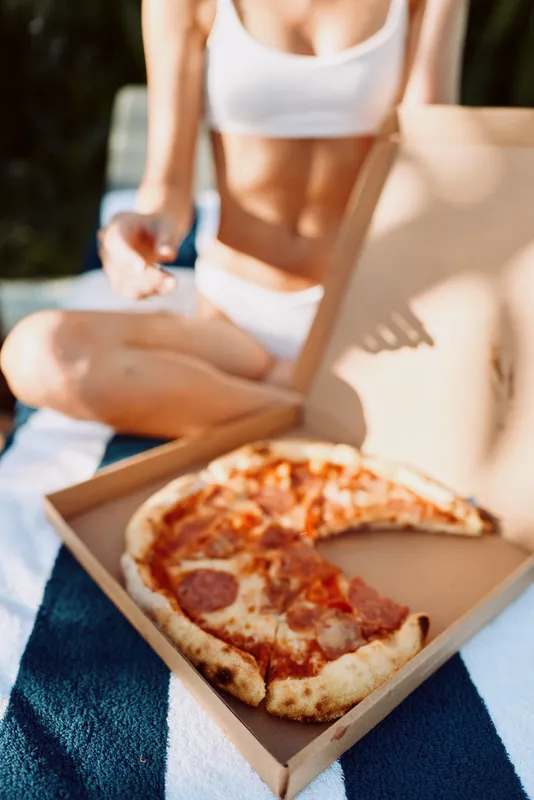 Casual outdoor lifestyle of a woman in a bikini enjoying pizza from a box on a towel by the beach, bright sunlight, relaxed mood, simple snack moment, candid and warm scene