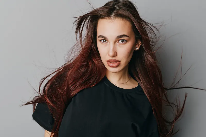 Young woman with long brown hair and black shirt posing confidently against gray background. Portrait with natural makeup and serious expression for fashion or beauty concept.