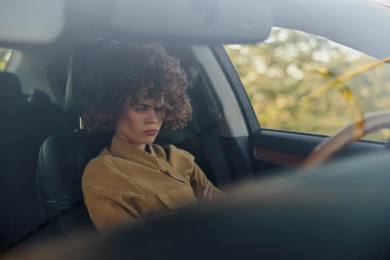 Woman sits in a car interior wearing a brown jacket, gazing downward. Soft retro film color tones create a warm, intimate lifestyle moment with a calm, reflective mood and everyday elegance.