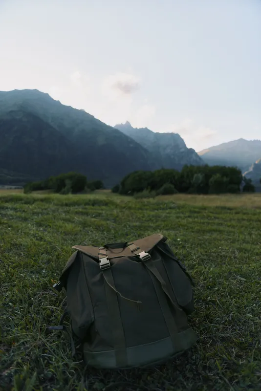 Backpack resting on a grassy field with distant mountains under a clear sky, outdoor adventure vibe, calm nature scene, ideal for hiking and travel moments