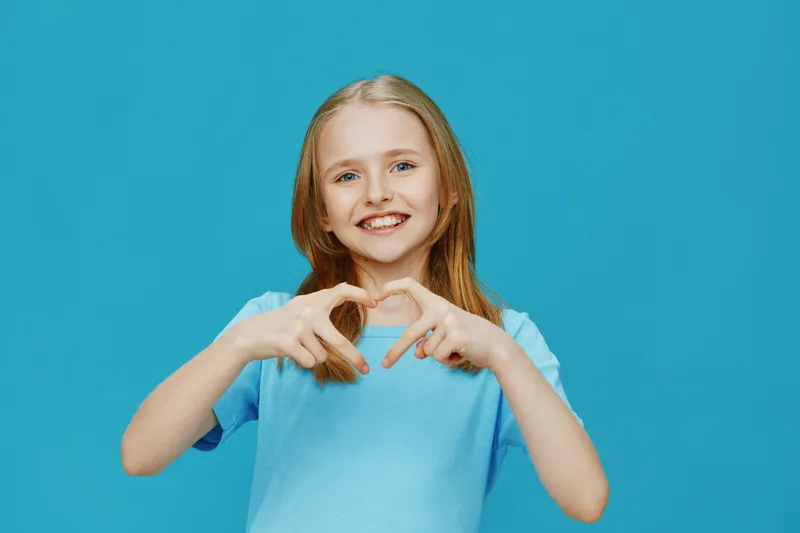Smiling girl in a blue t shirt making a heart shape with her hands against a bright blue background, representing joy and love, perfect for family and health themes