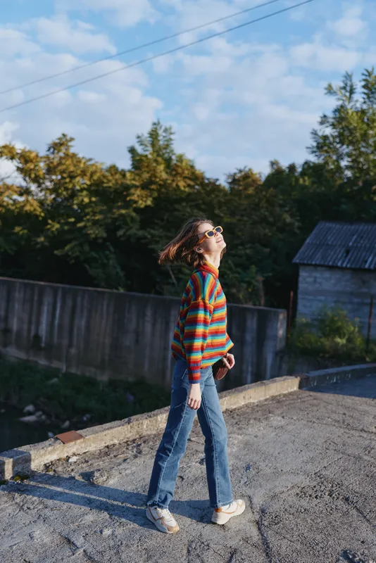 Woman walking on rooftop in colorful striped sweater and sunglasses, smiling and holding phone, young adult in casual jeans and sneakers, carefree lifestyle and sunny outdoor mood.