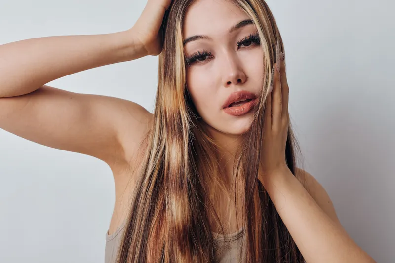 Woman with long light brown hair posing with hand on forehead and cheek, looking at camera with neutral expression. Studio portrait with natural makeup and smooth skin.