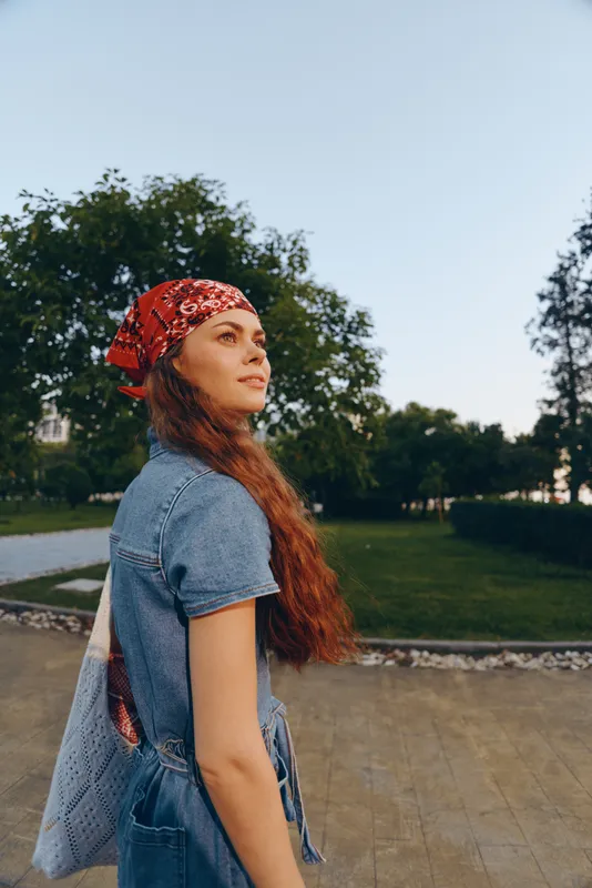 young woman with long hair wearing red headscarf and denim dress standing outdoors in park under clear blue sky during daytime