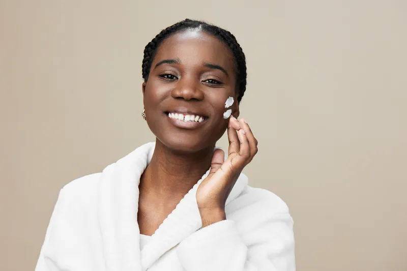 Beautiful young African woman applying a skincare cream, smiling with joy, wearing a cozy white robe, and set against a soft neutral background