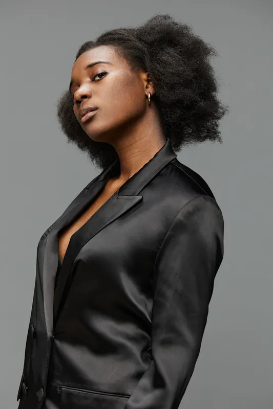 Elegant young Black woman in a sleek black blazer posing confidently against a minimalist gray background, showcasing style and empowerment
